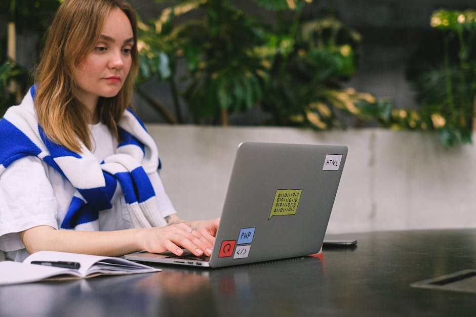 A young woman with shoulder-length light brown hair, wearing a white t-shirt and a blue and white striped scarf draped over her shoulders, is seated at a dark outdoor table. She is working on a silver